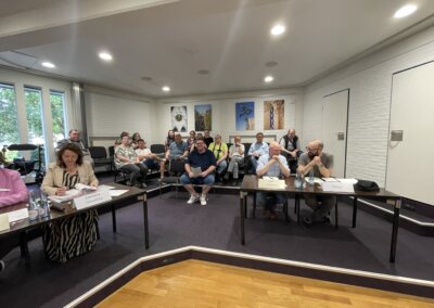 Eine Gruppe von Personen sitzt in einem Konferenzraum mit Blick nach vorne, mit U-förmig angeordneten Tischen und gerahmten Kunstwerken an den Wänden.