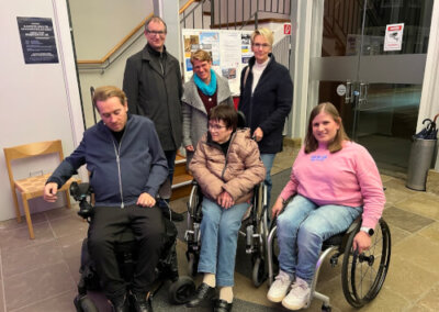 Sechs Personen, drei in Rollstühlen sitzend und drei hinter ihnen stehend, posieren für ein Gruppenfoto in einem Innenraum neben einer Schautafel und Glastüren.