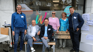 Sechs Personen posieren vor einem Schaufenster mit einem bunten W-Logo, stehen und sitzen auf einer Bank und lächeln in die Kamera.