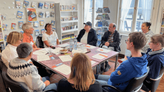 Acht Erwachsene sitzen in einer Bibliothek oder einem Besprechungsraum um einen Tisch herum und diskutieren; auf dem Tisch liegen Papiere, Bücher und Erfrischungen, im Hintergrund stehen Bücherregale.