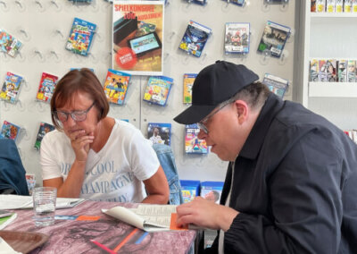 Zwei Personen sitzen an einem Tisch und lesen konzentriert, während an der Wand hinter ihnen Regale mit Videospielen und Zeitschriften hängen.