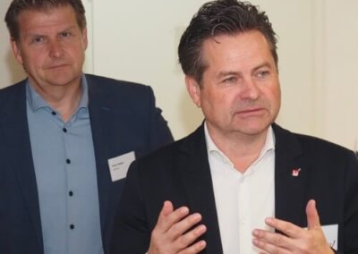 Zwei Männer in Geschäftskleidung stehen in einem Haus; der vordere Mann spricht und gestikuliert mit den Händen, während der andere Mann im Hintergrund steht und zusieht.