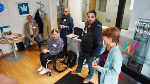 Fünf Erwachsene, darunter eine Frau im Rollstuhl, sind in einer kleinen Bürohalle versammelt. Einige sitzen, einige stehen, und sie scheinen sich zu unterhalten.