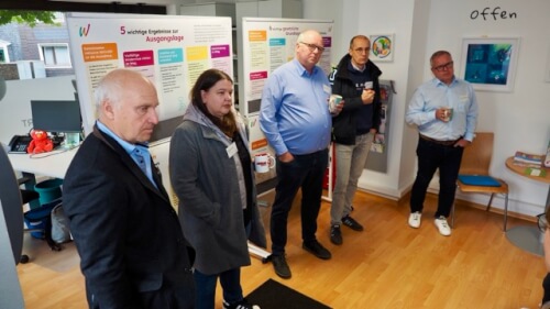 Fünf Personen stehen in einem Büro mit Informationsplakaten hinter ihnen, einige halten Tassen und hören aufmerksam zu.