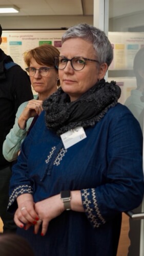 Zwei Frauen stehen in einem Haus, die eine vorne mit Brille, dunklem Schal und marineblau gemustertem Kleid mit Namensschild, die andere hinter ihr, ebenfalls mit Brille.