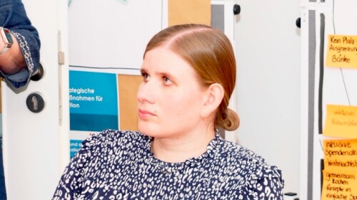 Eine Person mit zurückgebundenen hellbraunen Haaren sitzt in einem Raum und blickt nach links. Im Hintergrund sind Pinnwände mit Papieren und Notizen zu sehen.