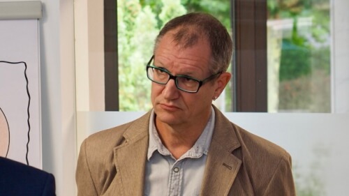 Ein Mann mit Brille und beigefarbenem Blazer steht in einem Haus und schaut mit neutraler Miene leicht zur Seite.