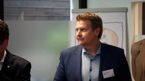 Ein Mann in einem blauen Anzug mit einem Namensschild steht drinnen neben einem Flipchart und schaut zur Seite.