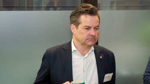 Ein Mann im Anzug, der einen Becher in der Hand hält, steht drinnen vor einer Milchglasscheibe, trägt ein Namensschild und blickt leicht nach unten.