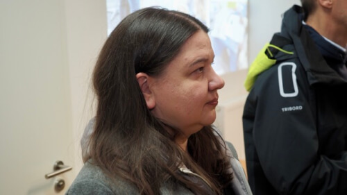 Eine Frau mit langen braunen Haaren und einem grauen Mantel steht in einem Haus und schaut nach rechts. Eine weitere Person in einer dunklen Jacke ist teilweise neben ihr zu sehen.