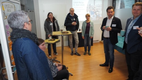 Eine Gruppe von acht Erwachsenen steht und sitzt in einem Raum mit Holzfußboden, einige mit Getränken und Snacks in der Hand, während einer gesellschaftlichen oder beruflichen Veranstaltung.