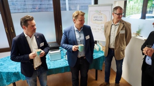 Vier Männer in Geschäftskleidung stehen und unterhalten sich an einem Tisch mit einem blauen Tuch und Kaffeetassen in einem scheinbaren Besprechungsraum eines Büros.