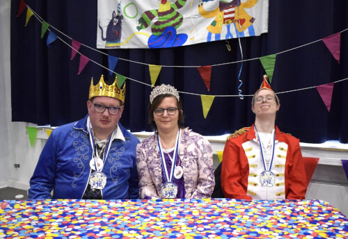 Drei Erwachsene, die Kronen und Medaillen tragen, sitzen an einem mit Konfetti bedeckten Tisch, mit festlichen Wimpeln und einem dekorativen Transparent im Hintergrund.