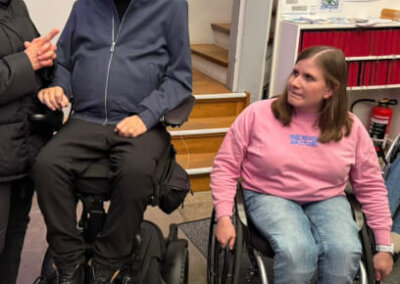 Ein Mann in einem motorisierten Rollstuhl und eine Frau in einem manuellen Rollstuhl unterhalten sich in einem Innenraum in der Nähe einer Treppe und einer Wand mit Postern und Informationsmaterial.