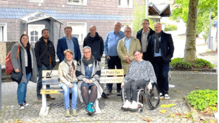 Eine Gruppe von elf Erwachsenen, darunter eine Person im Rollstuhl, posiert gemeinsam im Freien auf einem Kopfsteinpflasterweg in der Nähe von Bänken und Bäumen.