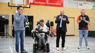 Vier Männer, von denen einer im Rollstuhl sitzt und eine Zeitung in der Hand hält, stehen während einer Veranstaltung in einer Sporthalle und sprechen in Mikrofone.