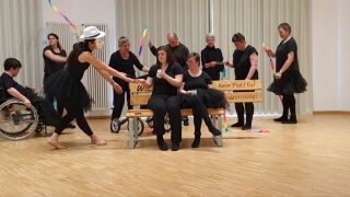 Eine Gruppe von Menschen, einige in schwarzen Kostümen und einer im Rollstuhl, tritt in einem hellen Raum um zwei Personen herum auf, die auf einer Holzbank mit der Aufschrift Kein Platz für Ausgrenzung! sitzen.