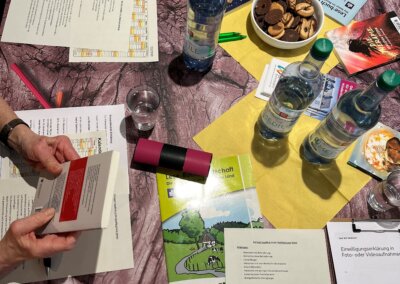 Ein Tisch mit Dokumenten, Broschüren, Wasserflaschen, Gläsern, einer Schale mit Keksen, Stiften und zwei Menschen, die Papiere in der Hand halten.