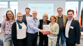 Acht Personen stehen in einem Innenraum zusammen, einige halten eine Urkunde oder eine Plakette in der Hand, im Hintergrund sind eine Präsentationswand und Fahnen zu sehen.