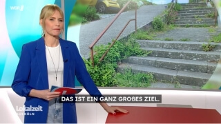 Eine Frau in einem blauen Blazer steht in einem Studio neben einem Bildschirm, auf dem ein Foto einer Außentreppe zu sehen ist; die deutschen Untertitel lauten: DAS IST EIN GANZ GROSSES ZIEL.