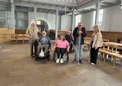 Sechs Erwachsene, davon zwei im Rollstuhl und einer mit einem weißen Stock, sitzen in einem geräumigen Raum mit Holzbänken und großen Fenstern.