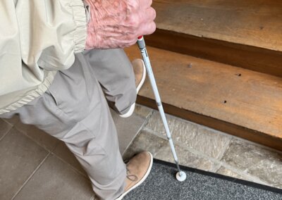 Eine ältere Person mit einem weißen Stock steht am Fuß einer Holztreppe und bereitet sich darauf vor, hinaufzugehen.