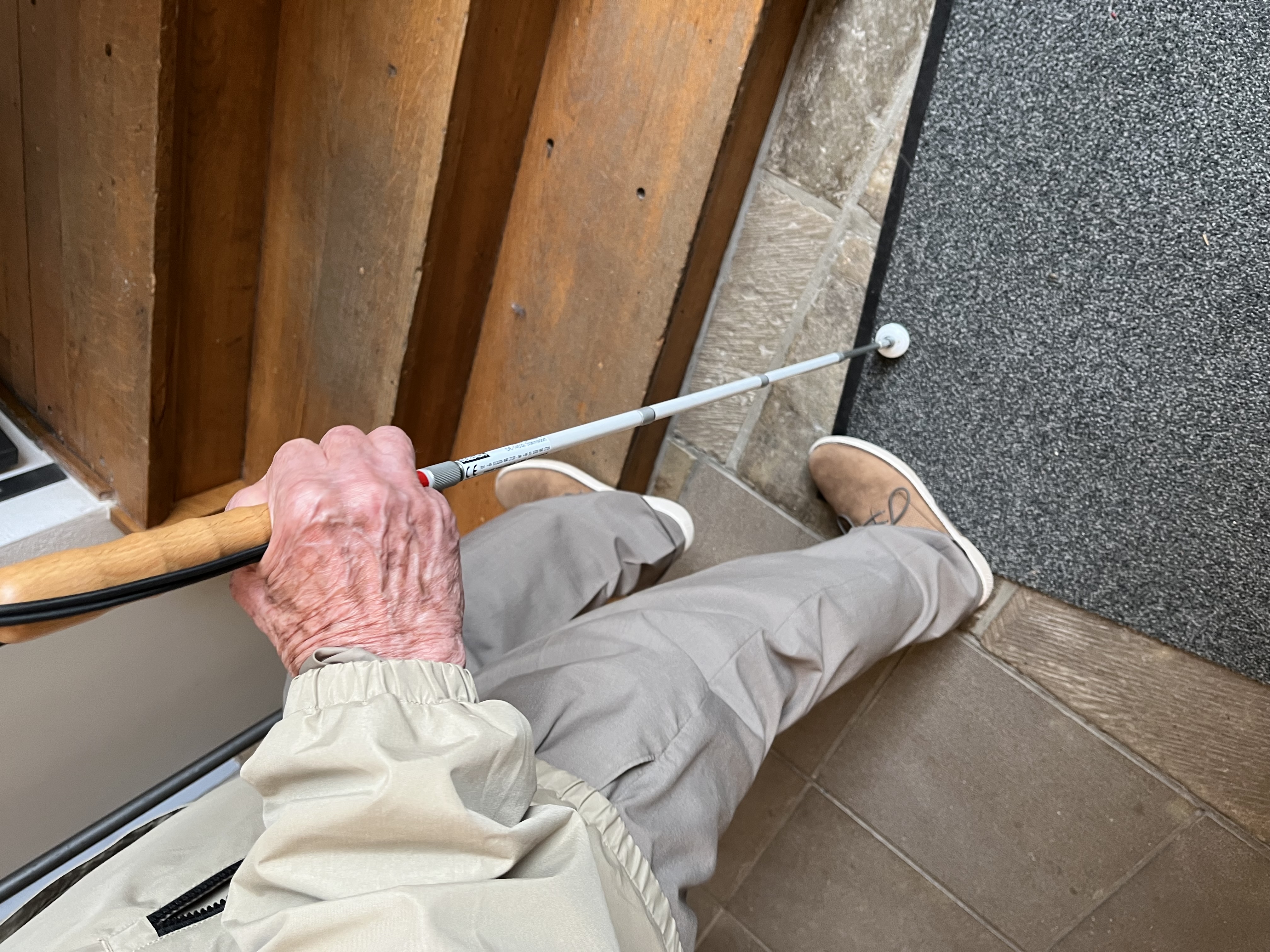 Eine ältere Person mit einem weißen Stock steht am Fuß einer Holztreppe und bereitet sich darauf vor, hinaufzugehen.