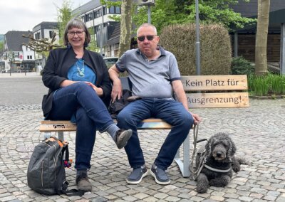 Zwei Personen sitzen auf einer Bank, auf der "Kein Platz für Ausgrenzung" steht, und neben ihnen liegt ein schwarzer Hund auf dem Boden.