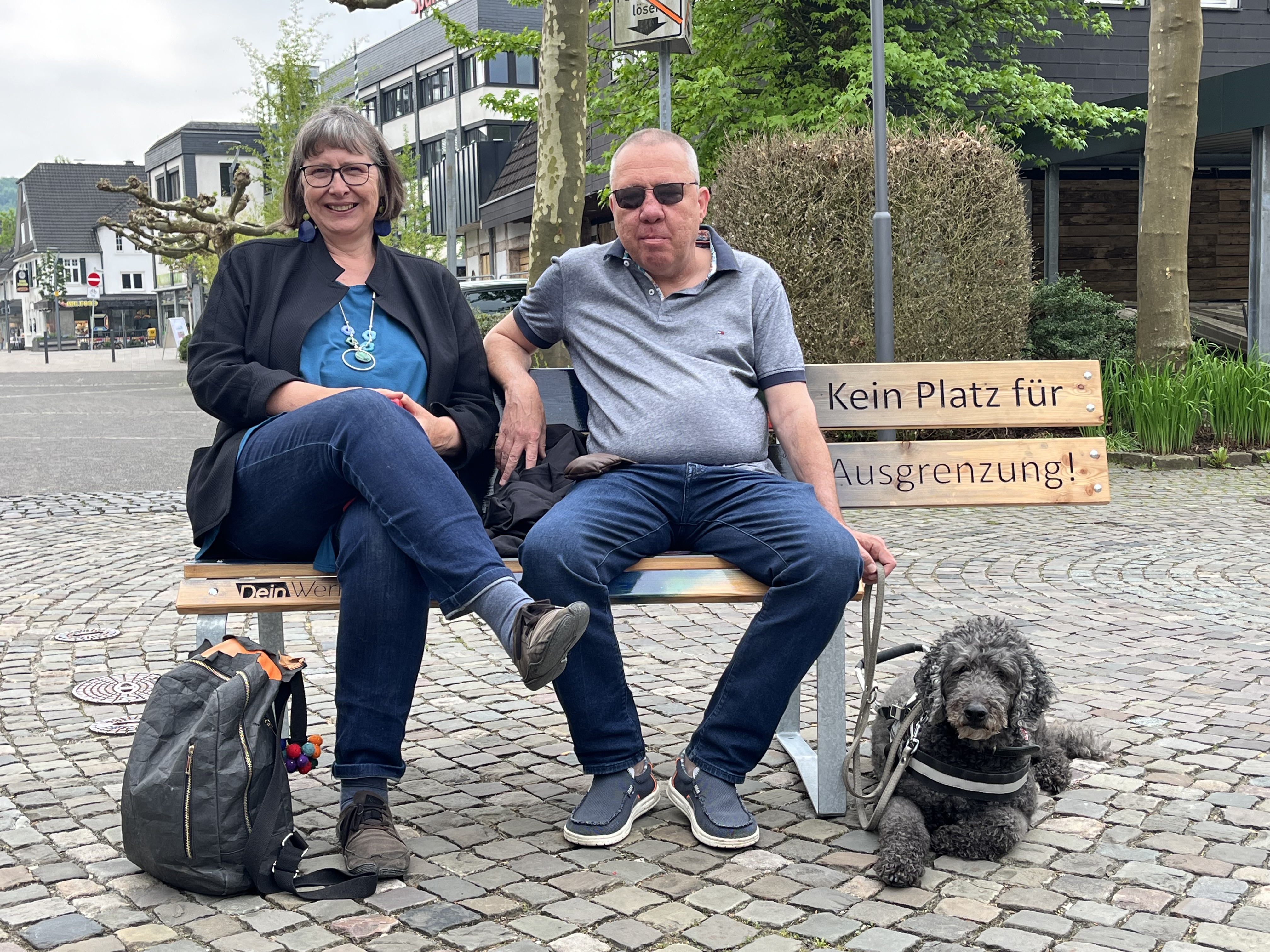 Zwei Personen sitzen auf einer Bank, auf der "Kein Platz für Ausgrenzung" steht, und neben ihnen liegt ein schwarzer Hund auf dem Boden.