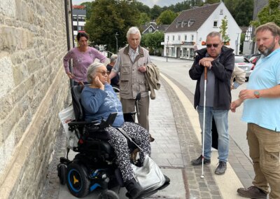 Fünf Erwachsene, darunter eine Frau im Rollstuhl und ein Mann mit einem weißen Stock, stehen an einem bewölkten Tag auf dem Bürgersteig neben einem steinernen Gebäude.