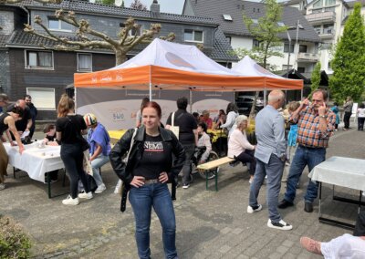 Menschen versammeln sich um Tische und unter einem orange-weißen Zelt bei einer Gemeindeveranstaltung im Freien; eine Frau steht im Vordergrund und schaut in die Kamera.