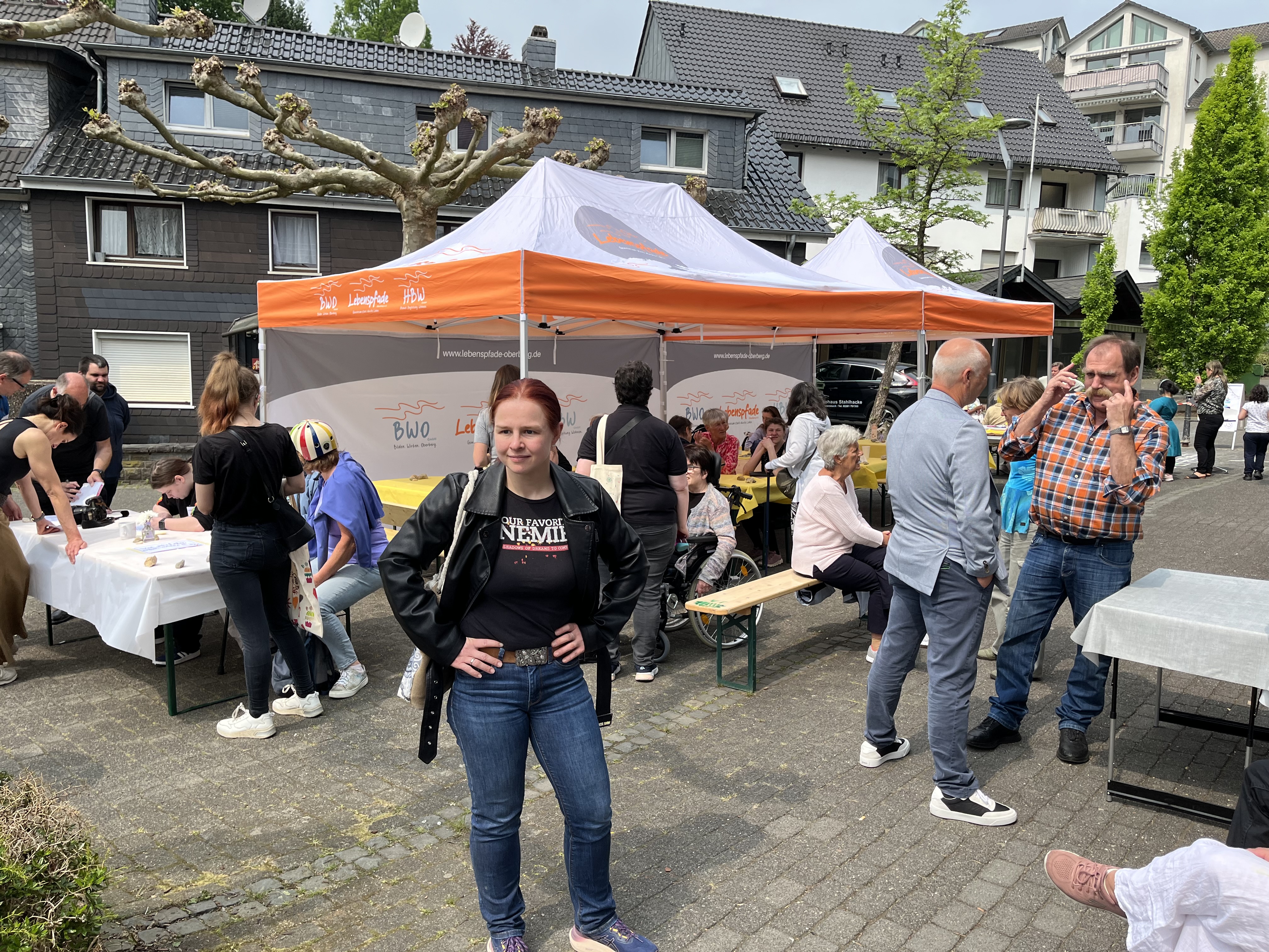 Menschen versammeln sich um Tische und unter einem orange-weißen Zelt bei einer Gemeindeveranstaltung im Freien; eine Frau steht im Vordergrund und schaut in die Kamera.