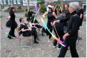 Eine Gruppe von schwarz gekleideten Personen tritt mit bunten Bändern in einem öffentlichen Außenbereich auf; eine Person trägt eine weiße Maske und einen Hut.