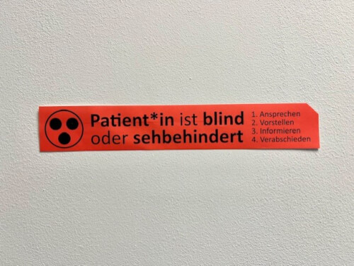 Rotes Schild an einer Wand mit deutschem Text, der angibt, dass der Patient blind oder sehbehindert ist, und einer Liste mit vier Schritten, die das Personal befolgen soll.