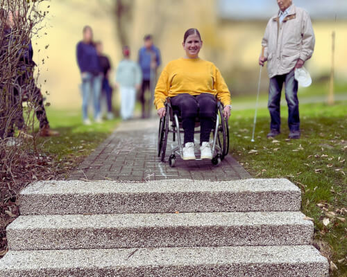 Eine Frau im Rollstuhl steht vor einer Treppe auf einem Weg im Freien, während im Hintergrund eine Gruppe von Menschen steht.