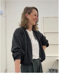 Eine Frau mit schulterlangen braunen Haaren, die ein weißes Hemd und eine dunkle Jacke trägt, steht in einem Raum und lächelt, während sie zur Seite schaut.