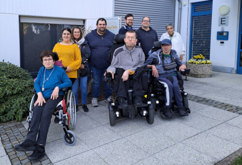 Eine Gruppe von neun Erwachsenen, darunter drei Rollstuhlfahrer, posiert gemeinsam vor einem Gebäude mit einer blauen Tür und Topfblumen.