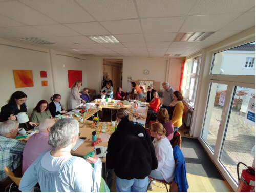 Eine Gruppe von Menschen sitzt und steht in einem hellen Raum um Tische herum, unterhält sich und beschäftigt sich, während auf den Tischen Essen und Getränke stehen.