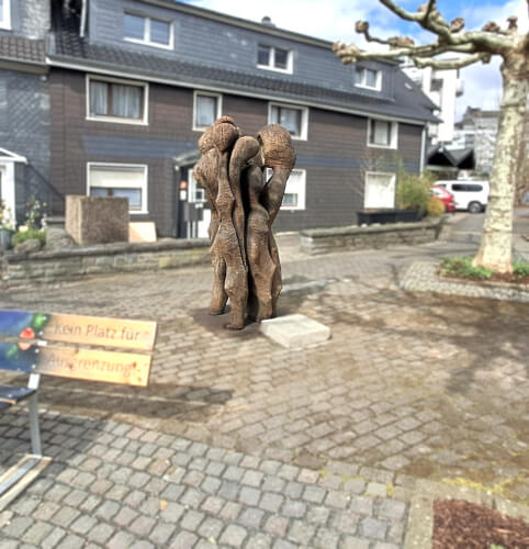 Eine Holzskulptur mit drei ineinander verschlungenen Personen steht in der Mitte eines gepflasterten Hofes vor Wohnhäusern.