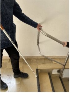Eine Person, die einen weißen Stock benutzt, nähert sich einer Treppe mit einem Handlauf, während eine andere Hand sich am Geländer festhält.