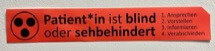 Rotes Etikett mit einem Symbol aus drei Punkten und einem Text in deutscher Sprache, der besagt, dass der Patient blind oder sehbehindert ist, mit der Anweisung zu sprechen, zu erklären, zu informieren und zu begleiten.