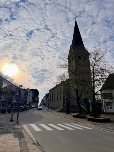 Eine Kirche mit einem hohen Kirchturm steht an einer ruhigen Straße unter einem teilweise bewölkten Himmel, durch den die Sonne scheint.