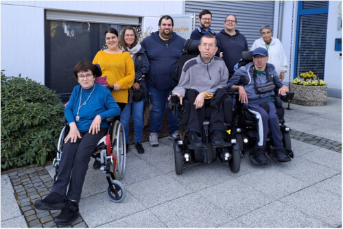 Eine Gruppe von neun Personen, darunter drei Rollstuhlfahrer, posiert für ein Foto vor einem Gebäude auf einer gepflasterten Fläche.