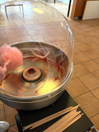 Eine Zuckerwattemaschine im Einsatz, die rosa Zucker auf einen Stock spinnt. Mehrere leere Holzstäbchen liegen auf dem Tisch daneben.