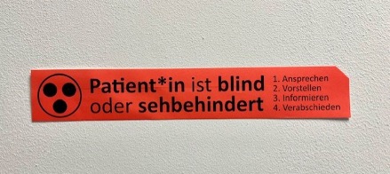 Rotes Schild in deutscher Sprache mit einem Symbol, auf dem steht: Der Patient ist blind oder sehbehindert und listet vier Kommunikationsschritte auf: ansprechen, vorstellen, informieren, verabschieden.