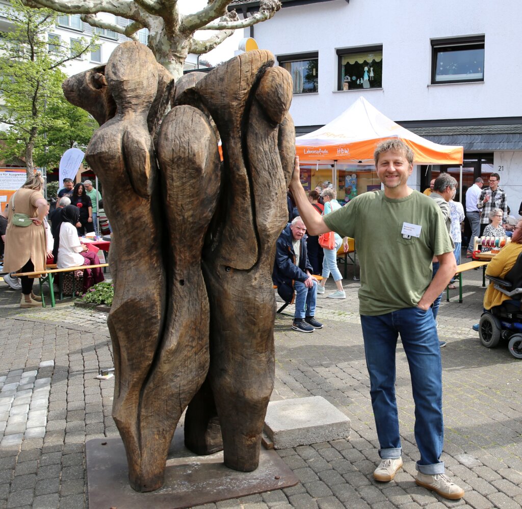 Ein Mann in einem grünen Hemd steht bei einer Veranstaltung im Freien neben einer großen abstrakten Holzskulptur mit Menschen und Marktständen im Hintergrund.