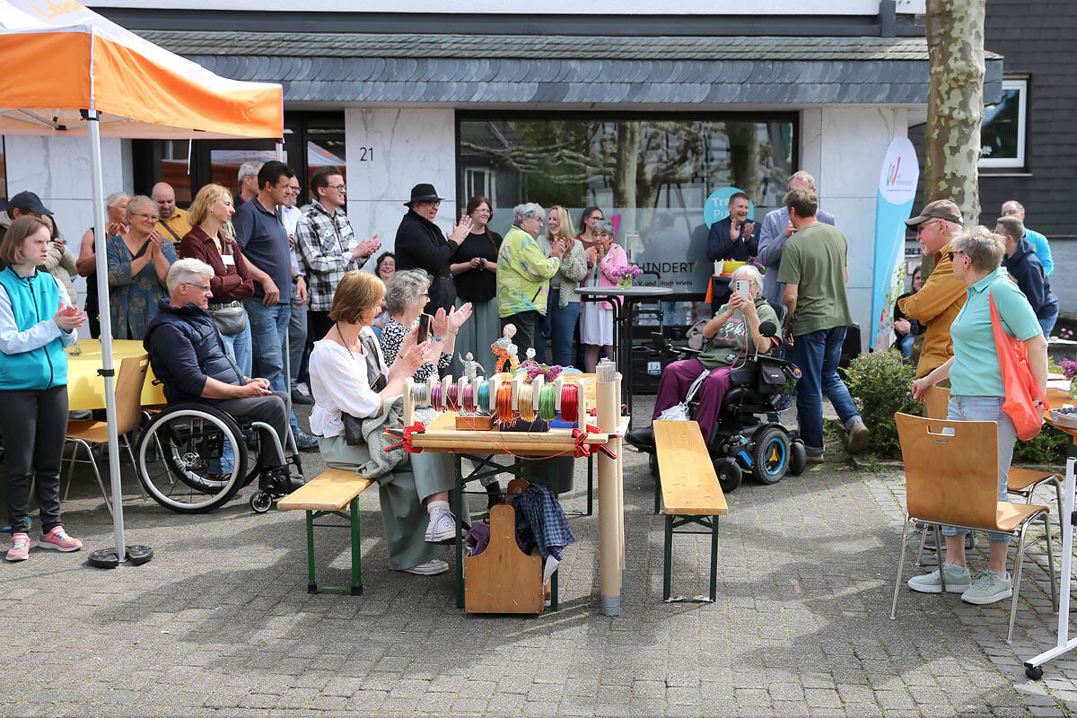 Eine Gruppe von Menschen, darunter mehrere im Rollstuhl, hat sich im Freien um eine Garnwickelstation versammelt und applaudiert, während eine Person vor einem Gebäude ins Mikrofon spricht.