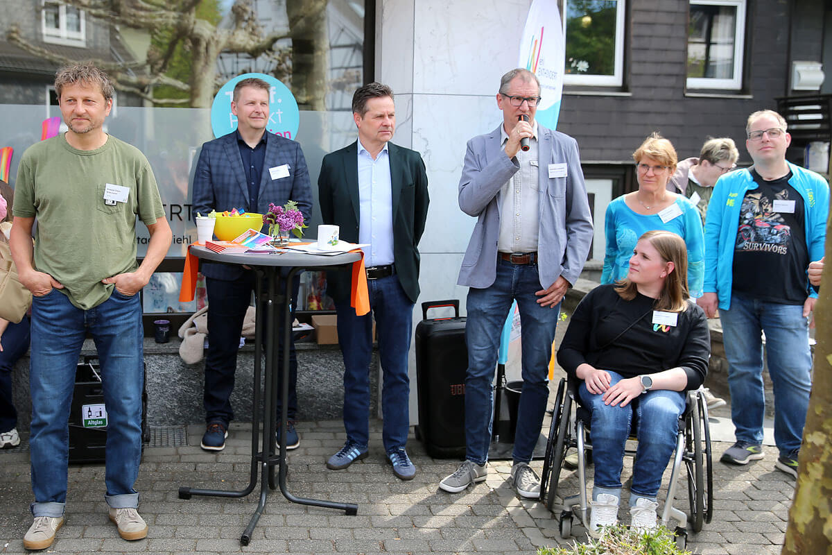 Eine Gruppe Erwachsener, darunter eine Person im Rollstuhl, steht im Freien in der Nähe eines Tisches mit Blumen und Getränken und hört zu; ein Mann spricht in ein Mikrofon.