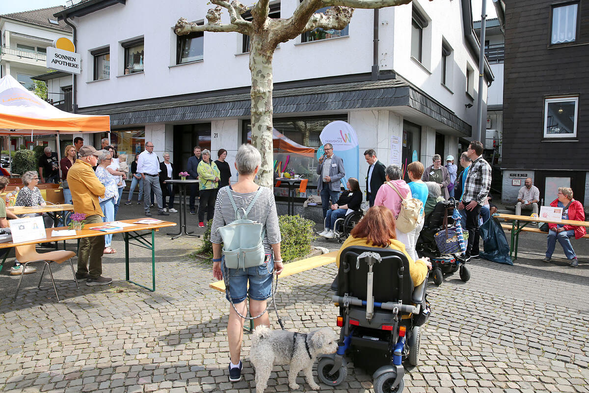 Die Menschen versammeln sich im Freien auf einem kleinen Platz mit Tischen, Zelten und Informationsständen. Einige sitzen, andere stehen, und eine Person sitzt im Rollstuhl, neben ihr ein Hund.