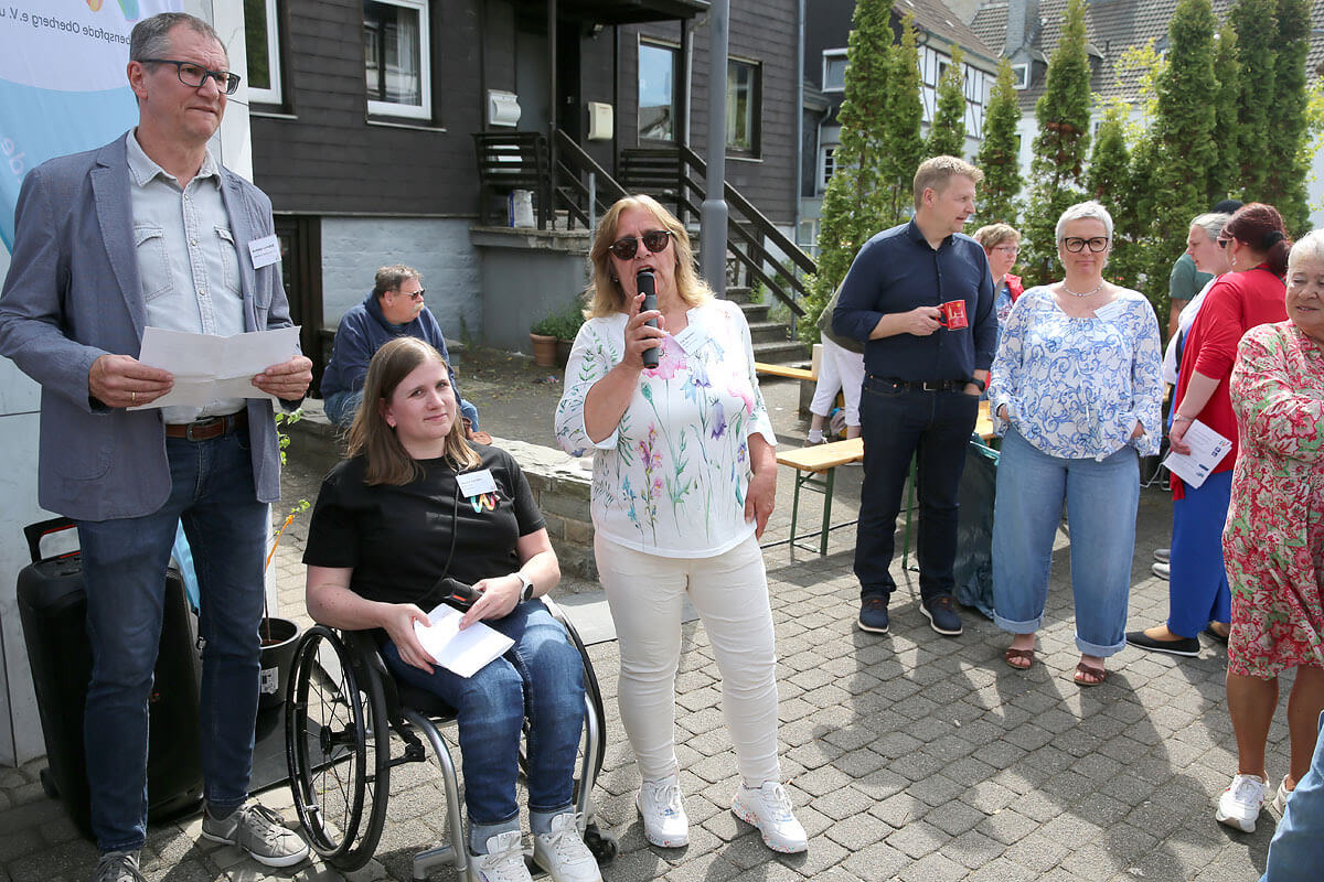 Eine Gruppe Erwachsener steht und sitzt im Freien auf einer gepflasterten Fläche; eine Frau spricht in ein Mikrofon, während die anderen zuhören.